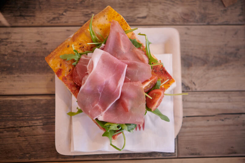 A square size of Sicilian pizza al taglio topped with prosciutto, arugula and cherry tomatoes