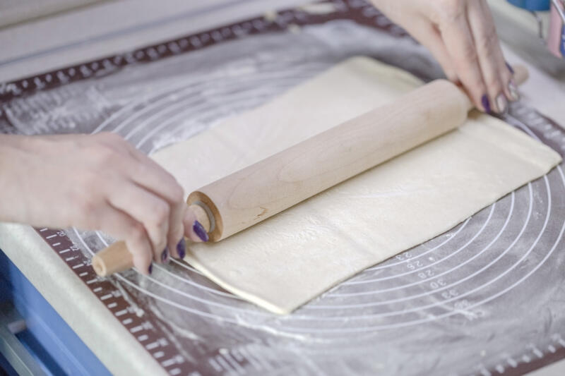 The woman rolls out a sheet of puff pastry with a wooden rolling pin on a silicone dough mat