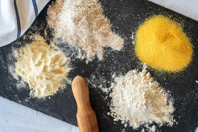 Different types of flour including white flour, whole wheat flour, durum wheat semolina, polenta flour, arranged on a slate cutting board