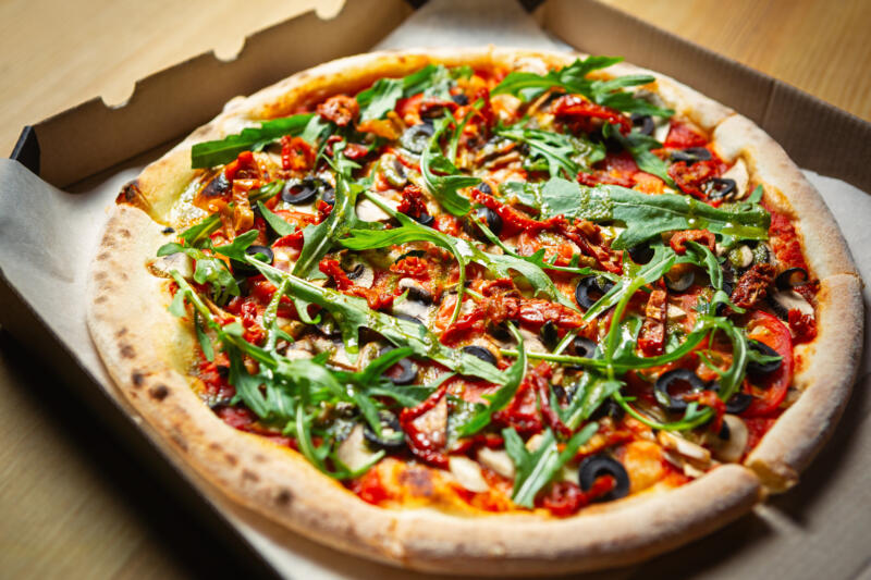 Italian style pizza with arugula, black olives, dried tomatoes, white champignon mushrooms in an opened pizza box