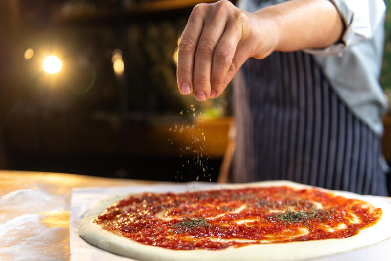 A human hand sprinkling a pizza with oregano
