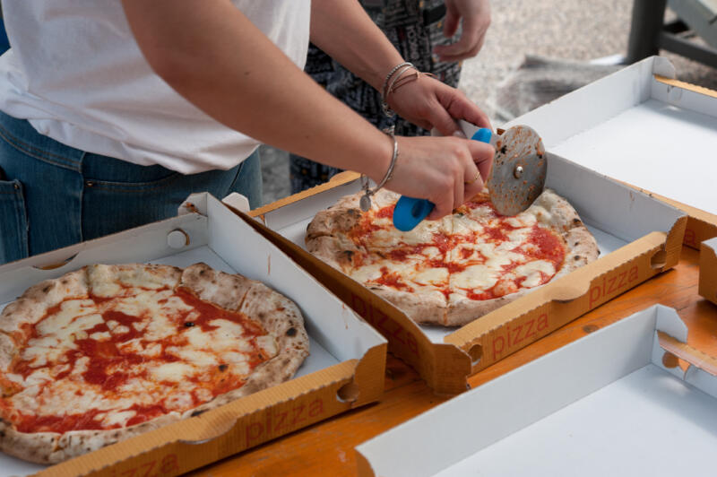 Margherita pizzas in pizza boxes for take away are being cut into slices by an employee
