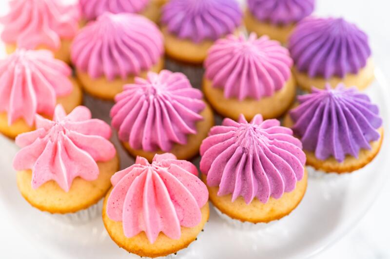 Freshly baked mini vanilla cupcakes with violet and pink buttercream frosting on a white cake stand