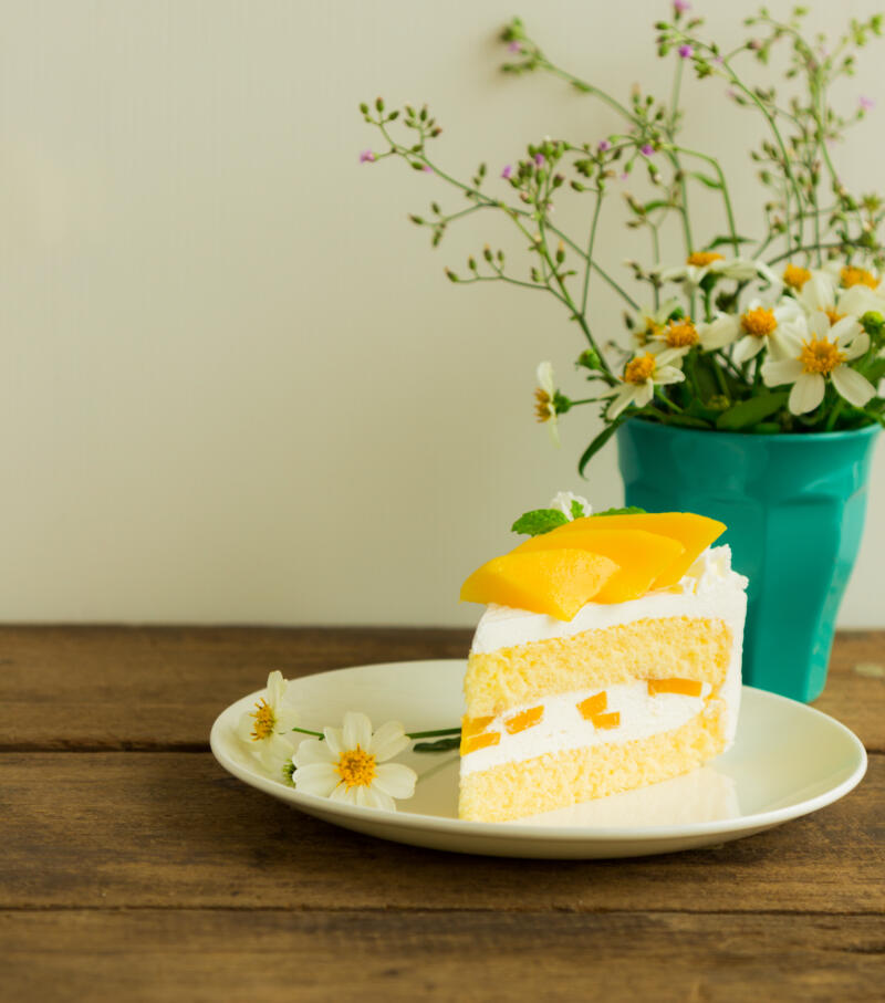 A slice of mango cake served on a plate on a wooden table decorated with a flowers in a vase
