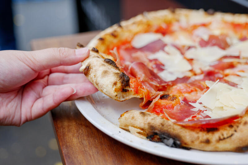 A hand holds a piece of Parma ham pizza from wood-fired oven