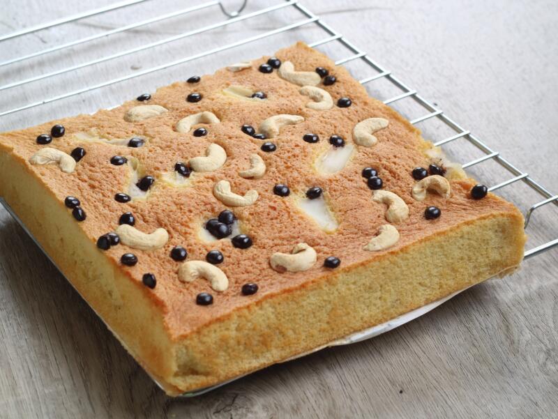 Square sponge cake topped with cashew nuts and chocolate cips on a rack to cool